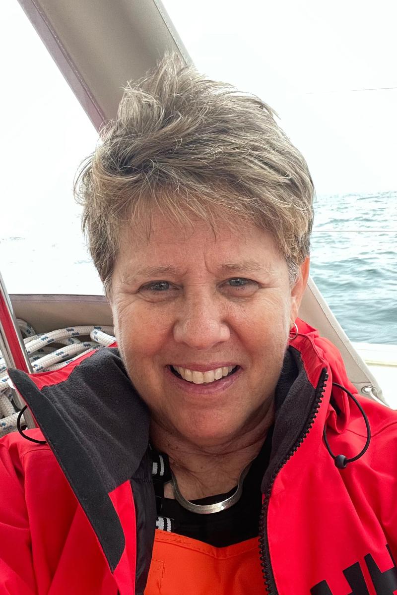 A smiling person wearing a bright red sailing jacket and orange foul-weather gear sits aboard a sailboat. Wind tousles their short hair as they face the camera. Coiled rope and parts of the boat are visible behind them, with open water stretching into the background under a bright, overcast sky.