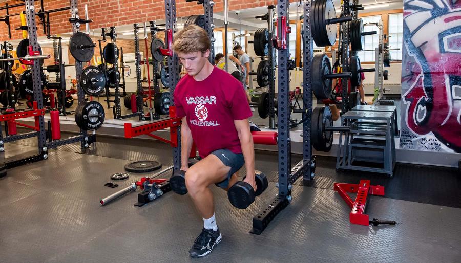 Male student wearing red shirt and gray shorts using hand weights in front of training equipment