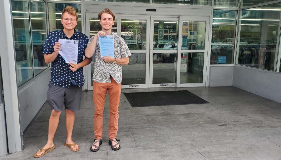 Christopher Unruh ’23 wearing navy blue patterned shirt, gray shorts and tan sandals and Chase Engel ’23 wearing gray and white patterned shirt, orange pants and brown sandals holding a document with immigrants’ legal rights in front of the doors of a  Goodwill store.