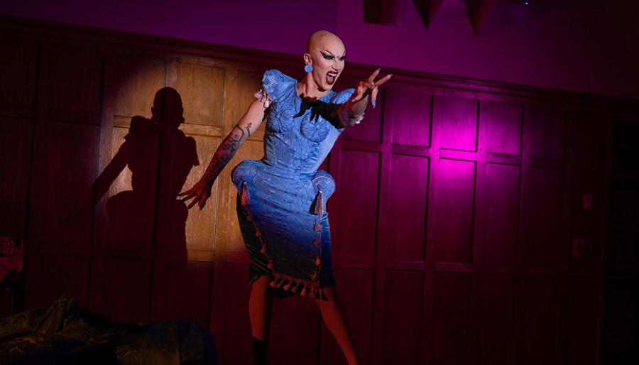 Woman in a blue dress and no hair singing on a dark stage.