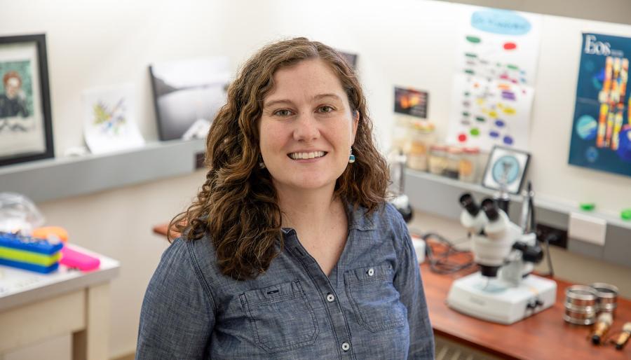 Person sitting on a stool smiling in a lab with scientific equipment in the background.