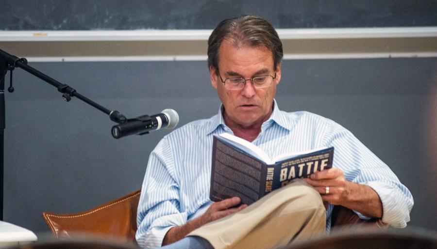 A person with glasses seated behind a microphone reading from a book with the work, Battle, and the cover.