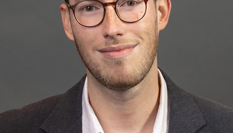 Person with brown short brown hair, tortoise shell glasses, white shirt with black blazer, smiling for a portrait. 