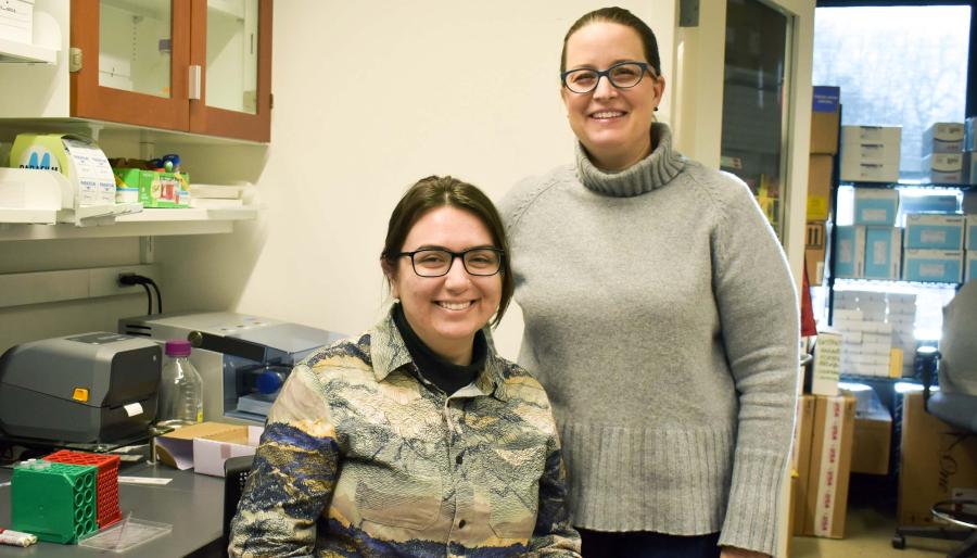 Morgan Stephens ’23 (left), sitting and wearing a printed collared shirt, and Visiting Scholar Elizabeth Thiele, standing and wearing a gray turtle neck sweater, in a research lab.