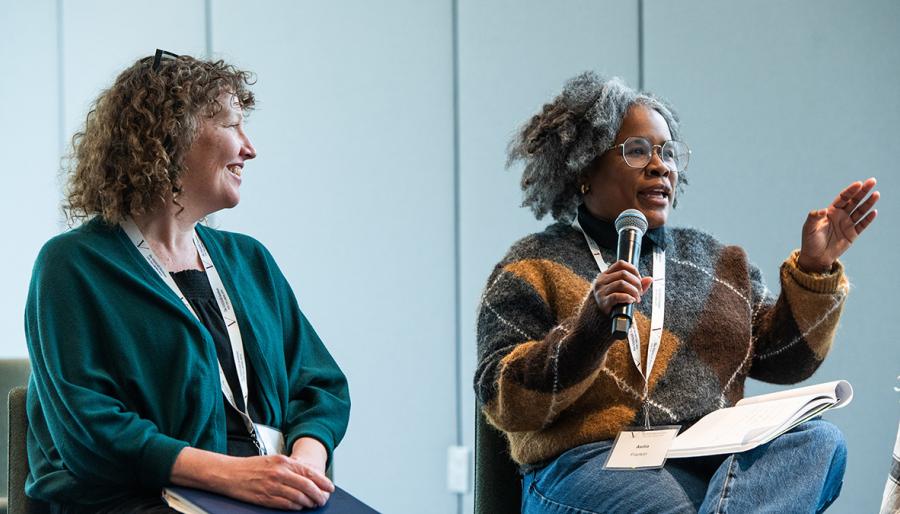 Two people sitting on a stage with one speaking into a microphone to an audience.