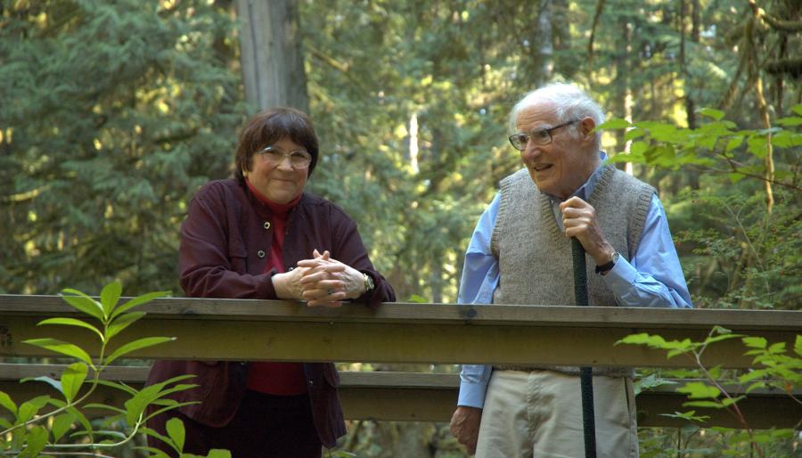 An elderly man and woman standing together on a wooden walkway in a lush, forested area, smiling and talking.