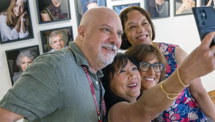 Four smiling taking a group "selfie" in front of a wall of portraits.