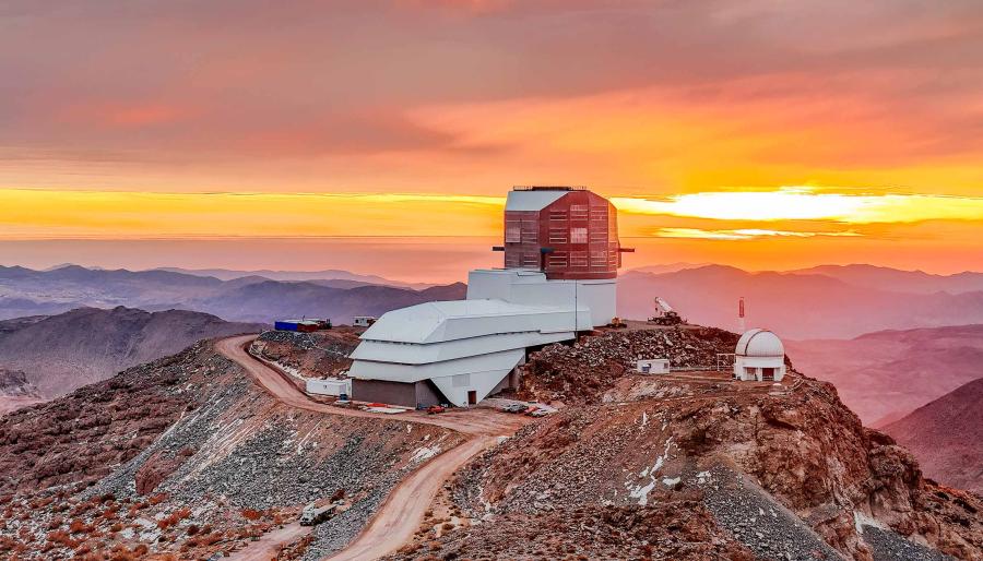 Vera C. Rubin Observatory sits atop a rocky mountain ridge at sunset, with vibrant orange and yellow hues lighting the sky.