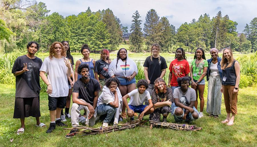 A diverse group of fifteen people is posing for a photo on a grassy lawn with a pond and a college building visible in the background.