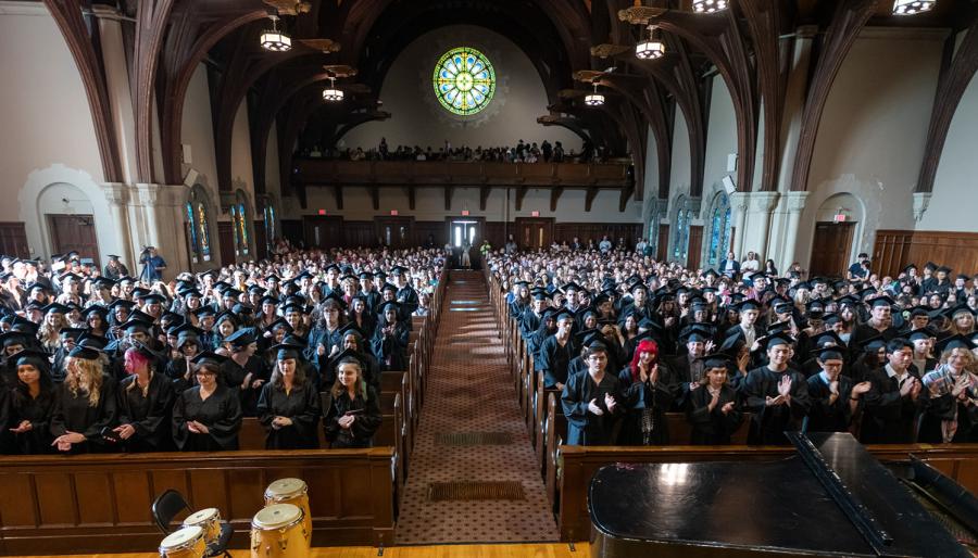 Large group of graduates in black caps and gowns filling a chapel, standing and applauding during a commencement ceremony, with stained glass windows and wooden beams visible overhead.
