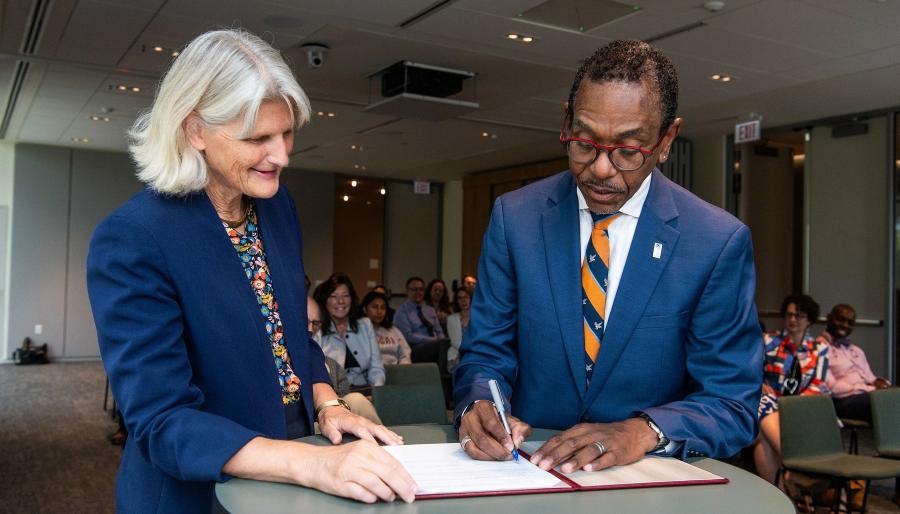 Two people signing a document at a table, with others seated in the background.