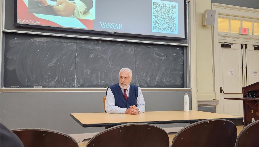 Person seated at a classroom table in front of a chalkboard, with a Vassar presentation slide and QR code projected above.