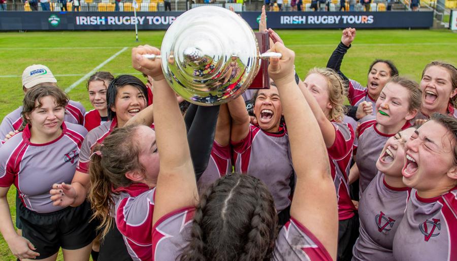 Vassar College Women’s rugby team in maroon and gray jerseys celebrating on the field, holding up a large silver trophy together with excitement.