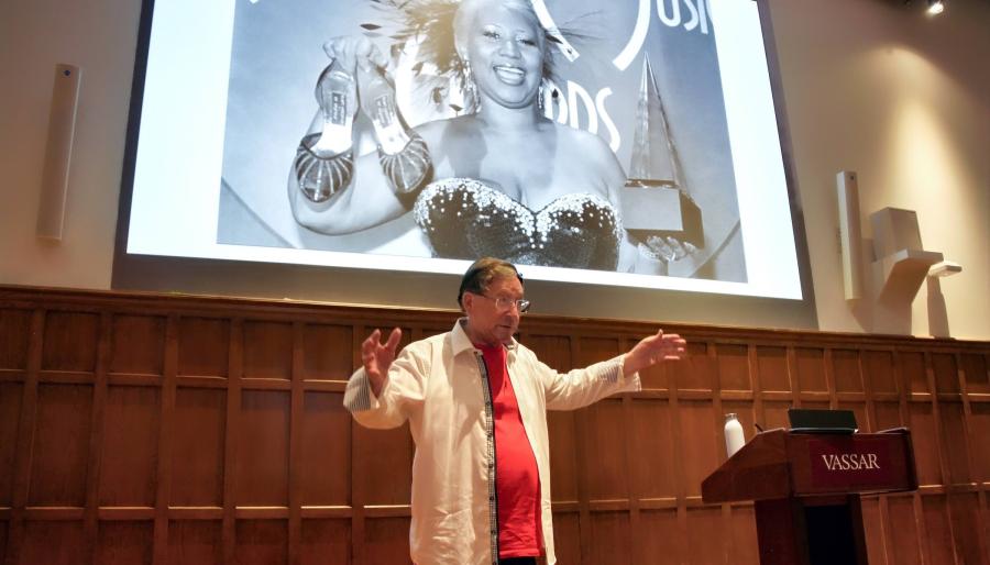 A person standing and speaking before and audience and in front of a large projection screen with their arms spread wide.