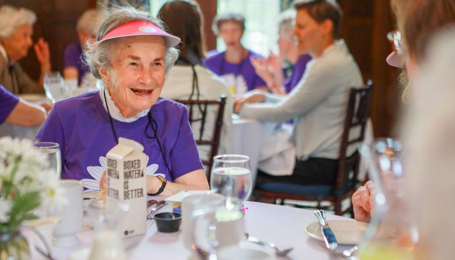 June Ross Marks ’49 with short white hair wearing a bright purple shirt and a pink visor, seated at a dining table.