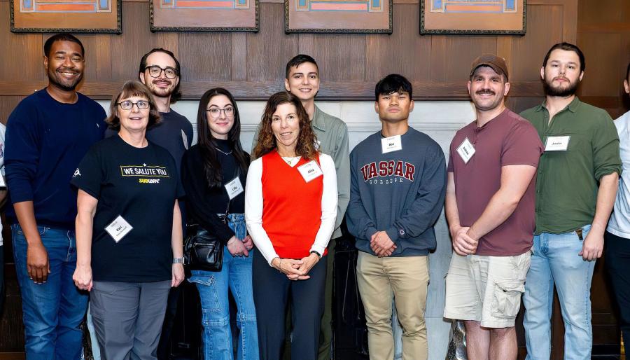 Eleven people stand in a line indoors, smiling at the camera, wearing name badges; one in a “We Salute You” T-shirt, one in a Vassar sweatshirt, one in a red sweater, with wood-paneled walls and framed art behind them.