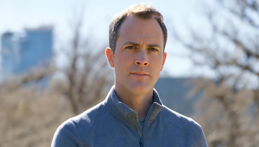 Outdoor head-and-shoulders portrait of a person in a gray quarter-zip, neutral expression, with blurred trees and a blue building behind the person.