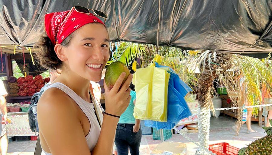 Smiling shopper in a red bandana and sunglasses holds a mango at an outdoor market, standing under a tarp beside hanging yellow and blue bags, with produce stalls and palm fronds in the background.