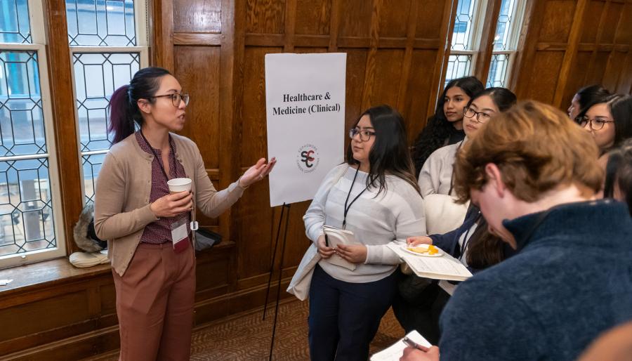 A speaker gestures while addressing a small group of students wearing name badges at a networking event near a “Healthcare & Medicine (Clinical)” sign.