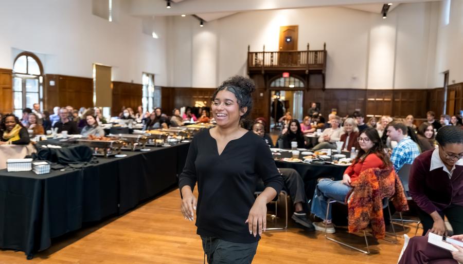 A smiling attendee walks through a large hall as seated guests watch, with tables of food and a wood-paneled room in the background.
