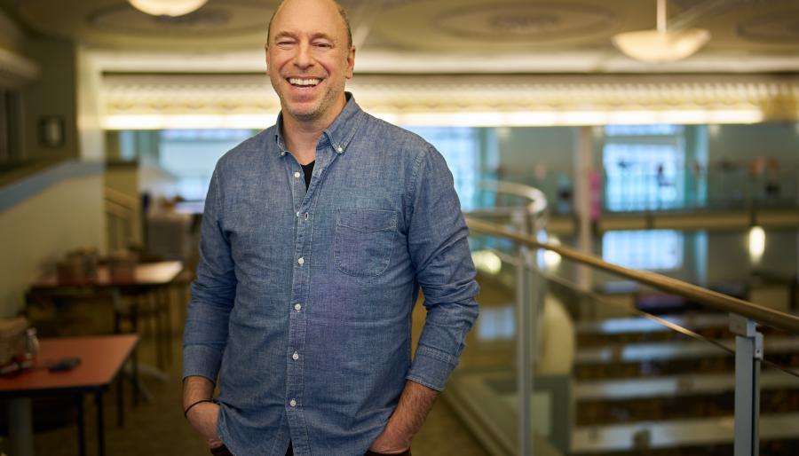 A smiling person wearing a denim shirt stands with their hands in their pants pockets in an indoor atrium with big windows.