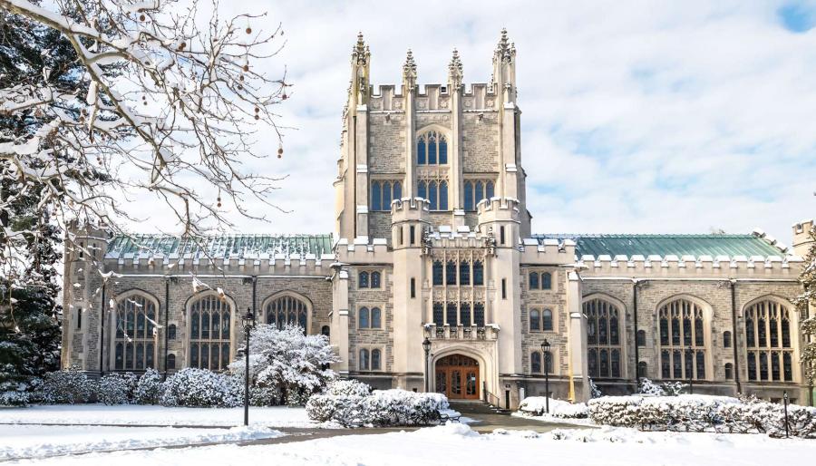 Winter view of a Gothic-style stone campus building with a tall central tower, arched windows, and a green roof, surrounded by snow-covered trees and shrubs under a partly cloudy sky.