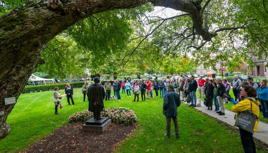 Group of visitors gathered on a campus lawn around a statue during an outdoor tour, with large tree branches overhead.
