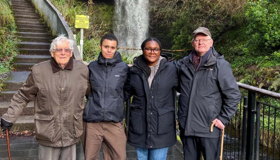 Four people stand outdoors, with foliage and a stone staircase behind them.