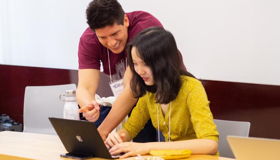 Two conference participants collaborate at a table, with one person pointing at a laptop screen while the other works on the computer. Both wear conference name badges, and reusable water bottles and laptops are visible in the bright classroom setting, suggesting a hands-on workshop or collaborative learning session.