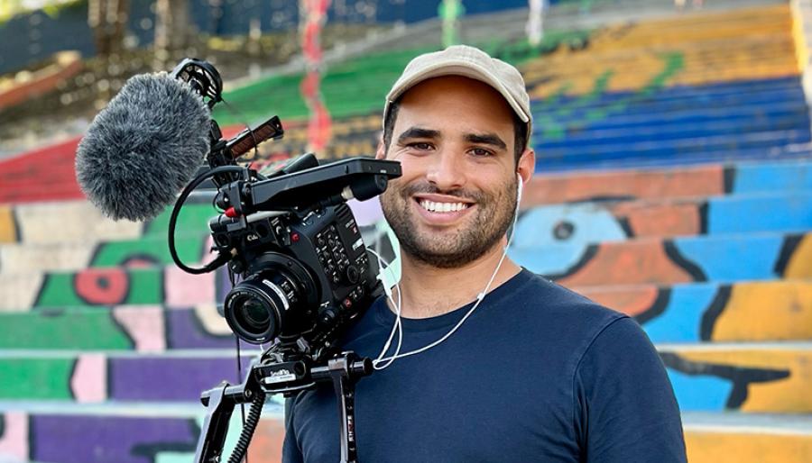 A smiling man stands beside a video camera and microphone, with a colorful mural behind him.