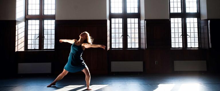 A person stands in a large space with tall windows, posing in the sunlight. 