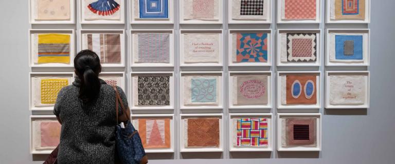 A person stands with their back to the viewer, looking at a grid of colored artworks on a gallery wall.