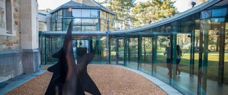 A walkway enclosed in glass windows curves past a metal sculpture. Trees are visible in the background.