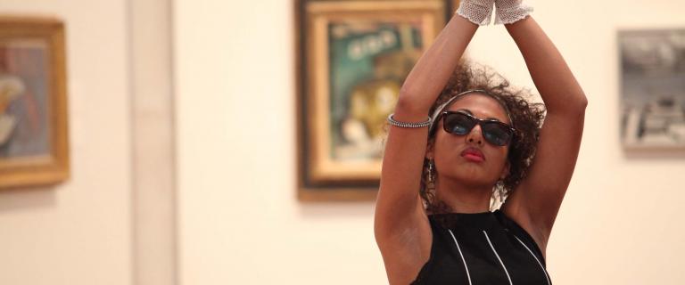 A person with sunglasses and black curly hair poses in front of paintings in a gallery.