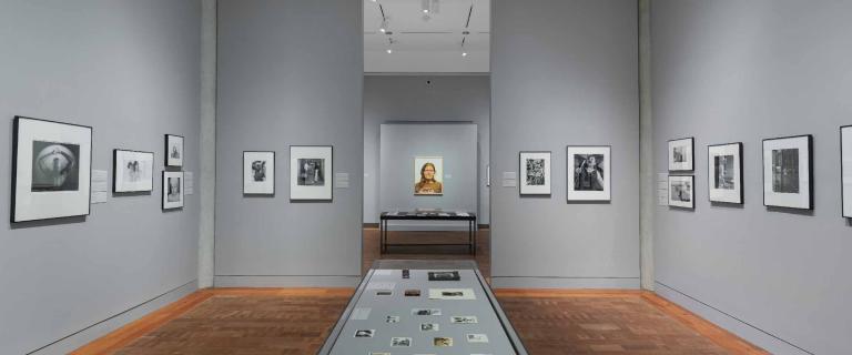 The interior of a gallery: gray walls, monochrome photos on the walls, and a table with more photos in the middle of the room.