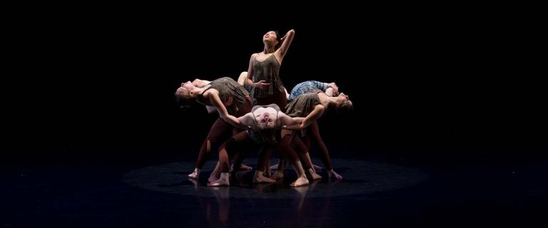 Several dancers pose on a darkened stage. All except one are bending backwards; one stands in the middle, looking upward.