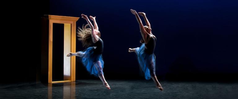 Two people wearing dresses leap across a darkened stage, arms in the air.