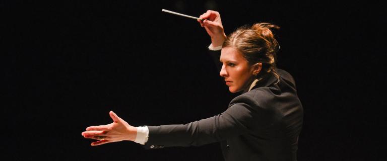 Dramatically lit side view of Lidiya Yankovskaya ’08, alone, holding a baton while directing an orchestra offscreen