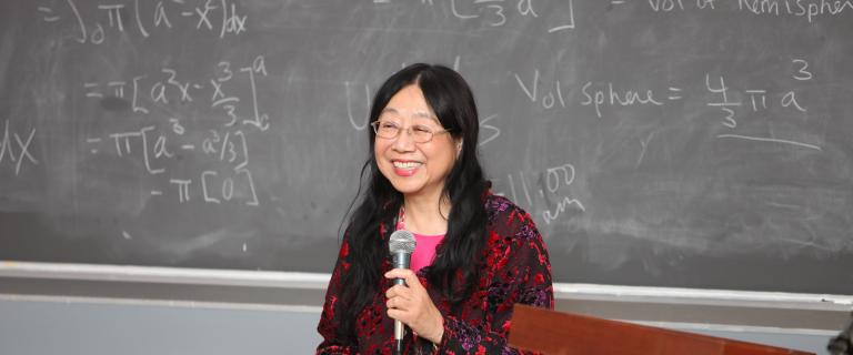 Physicist Sau Lan Wu ’63 speaking in front of a chalkboard with a microphone