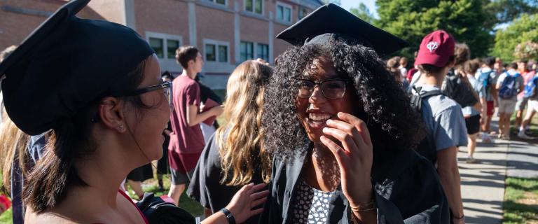 Two students wearing black caps and gowns laughing and other students in the background at Vassar College Convocation.