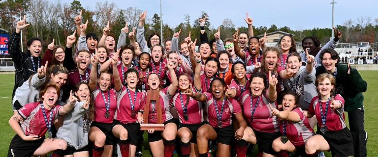 Group shot of Vassar women's rugby team on the field holding up hands making the number one sign