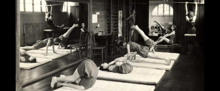 Vassar’s Calestheneum opened in 1866 showing women in old style gym suits working out on white mats and a hanging bar in front of a large mirror 