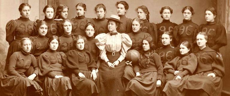 Group black and white photo of Vassar’s 1896 women's basketball team wearing black long skirt and long puffy sleeved shirts with the number 96 on them