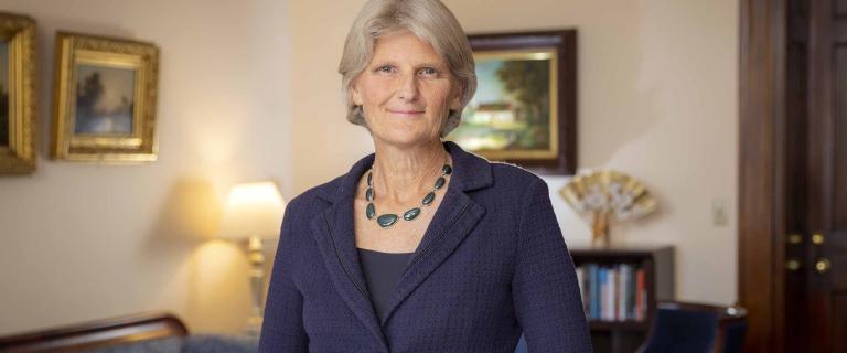 A person with straight gray hair, a necklace of shiny black stones, and an indigo jacket smiles at the camera
