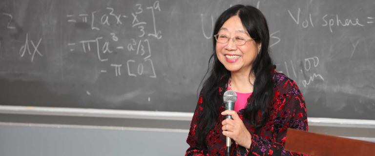person in front of a chalk board holding a microphone smiling and speaking to an audience off camera