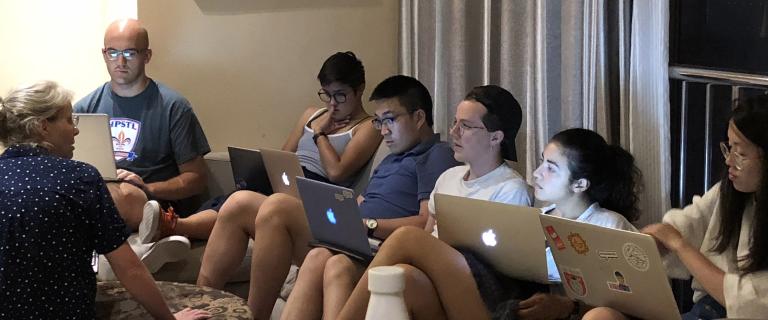 Students sitting in a hotel room lined up agains the wall with Apply laptops resting on their laps.
