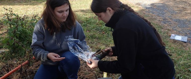 Two students outside kneeling on the ground bagging soil samples in a plastic bag.