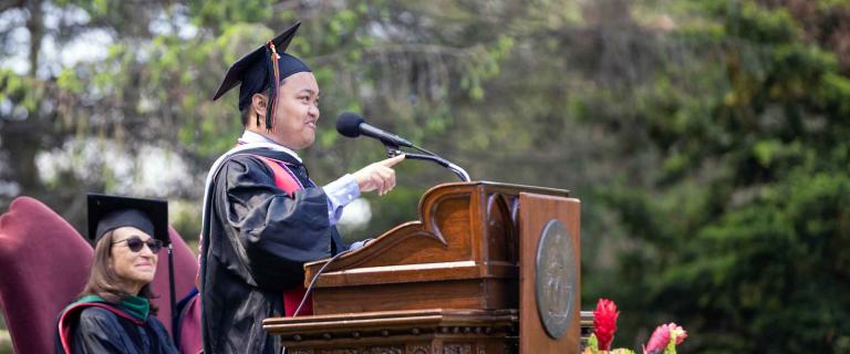 Commencement speaker speaking and pointing behind a podium and microphone with faculty and administrators watching