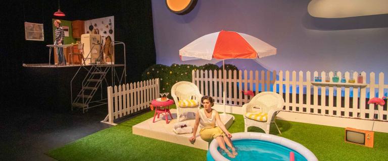 two actors on a stage with a backyard pool set with an umbrella and a kitchen