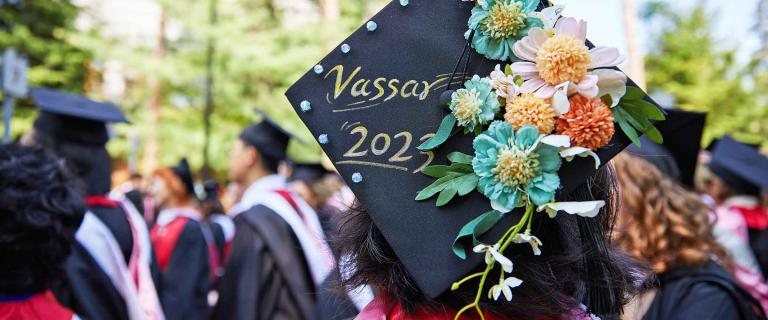 A close-up photo of a graduation cap with flowers on it and ornate hand-written letters that say "Vassar 2023". The person wearing it stands outside in a crowd of other graduates.